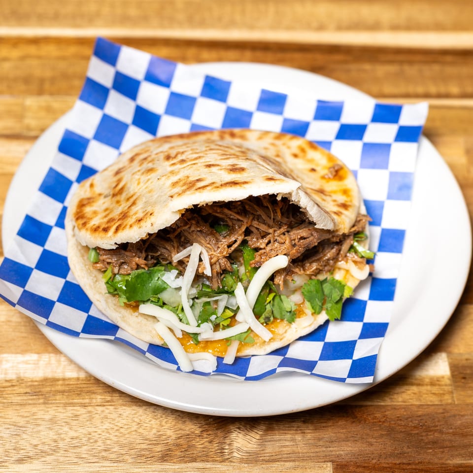A pita bread sandwich filled with shredded meat, cilantro, onions, and cheese on a white plate.