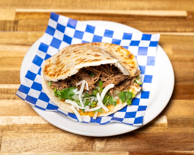 A pita bread sandwich filled with shredded meat, cilantro, onions, and cheese on a white plate.