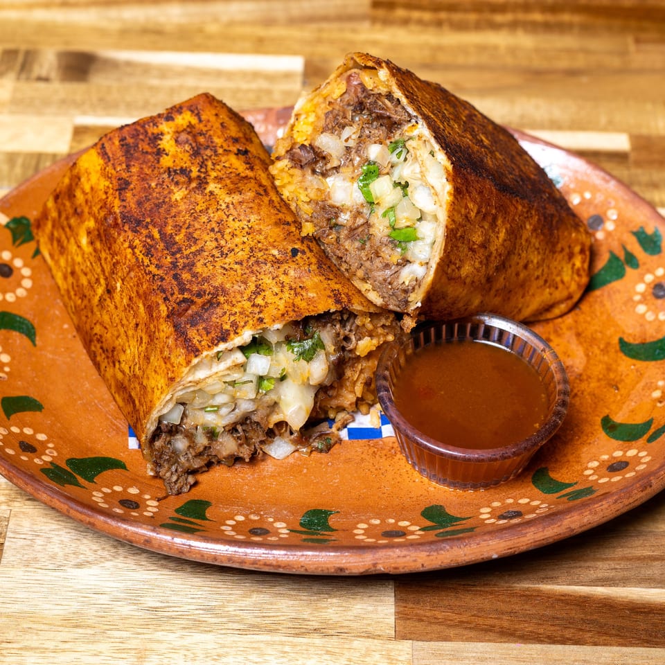 Two halves of a large, grilled burrito filled with meat, rice, and onions, served with dipping sauce.