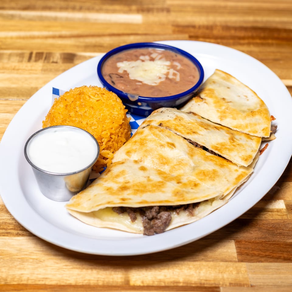 A white plate with beef quesadillas, rice, refried beans, and sour cream on a wooden table.