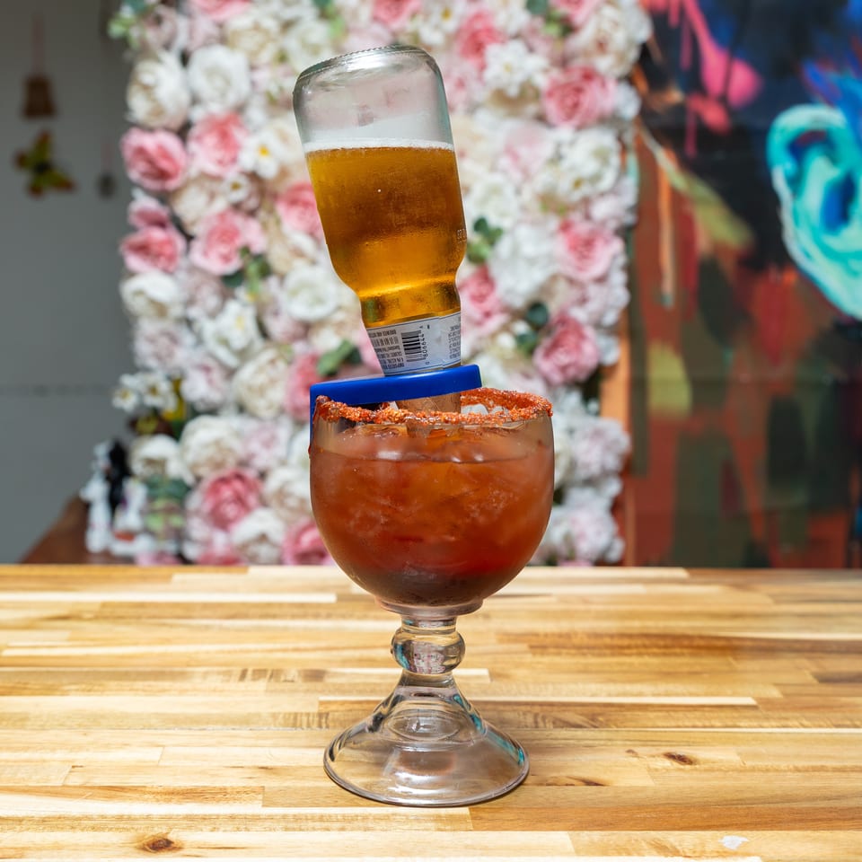A michelada with a beer bottle inverted into it, on a wooden table with floral decor.