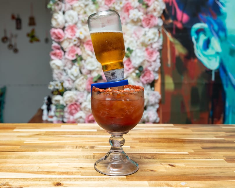 A michelada with a beer bottle inverted into it, on a wooden table with floral decor.