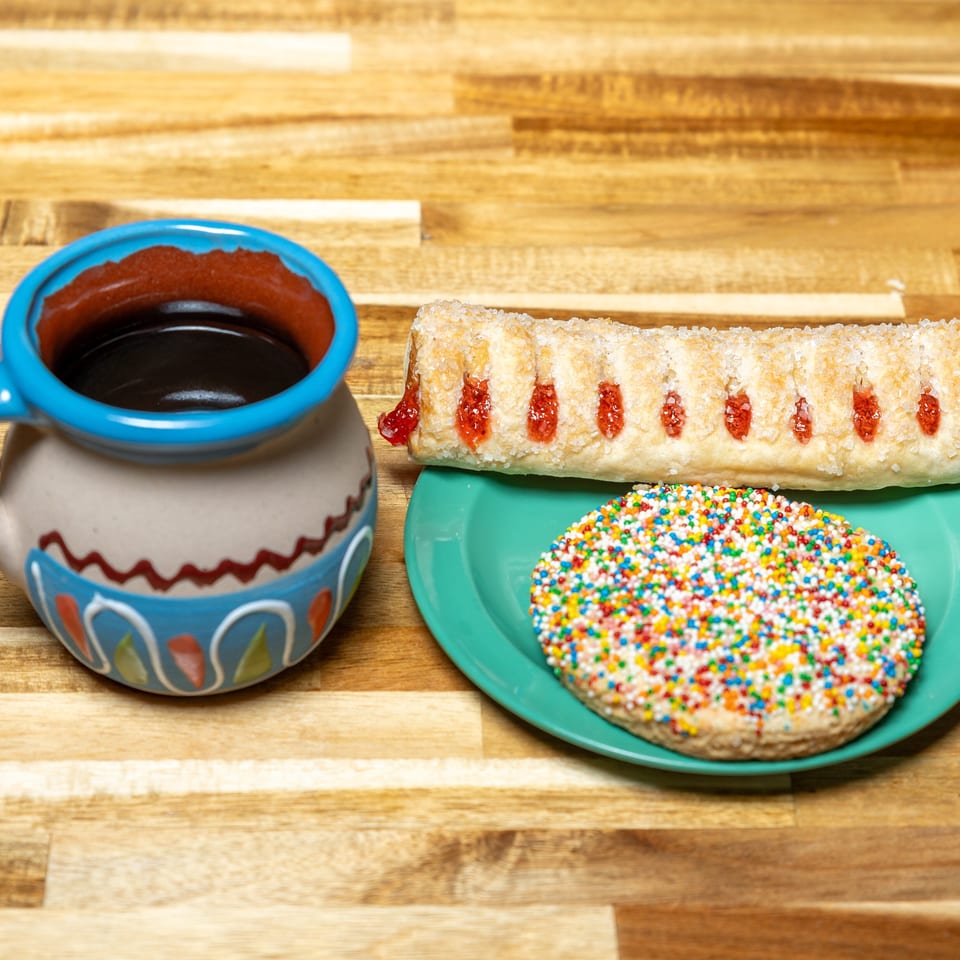 A colorful mug of coffee sits next to a plate with a sprinkle cookie and a guava pastry.