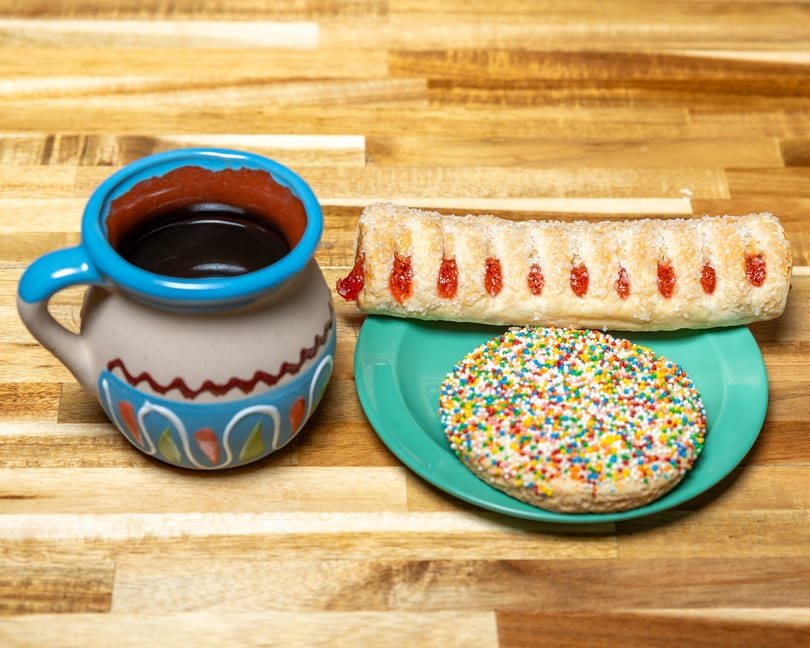 A colorful mug of coffee sits next to a plate with a sprinkle cookie and a guava pastry.