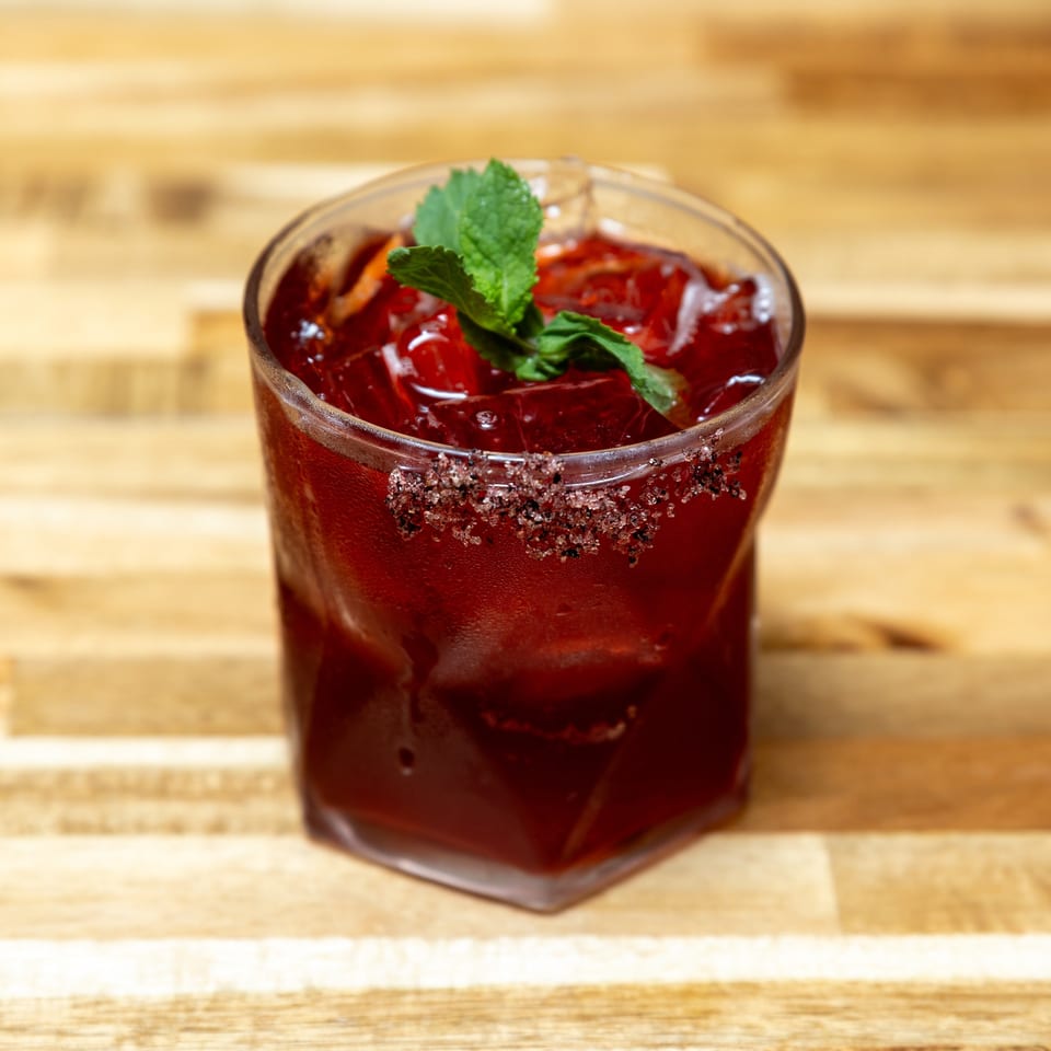 A vibrant red cocktail with ice, garnished with mint and a sugar-rimmed glass on a wooden surface.