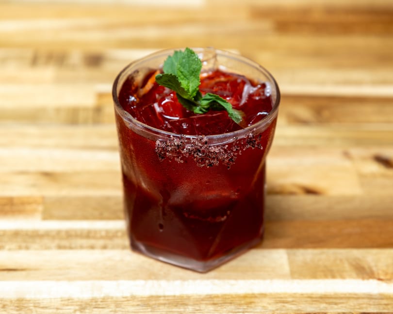 A vibrant red cocktail with ice, garnished with mint and a sugar-rimmed glass on a wooden surface.