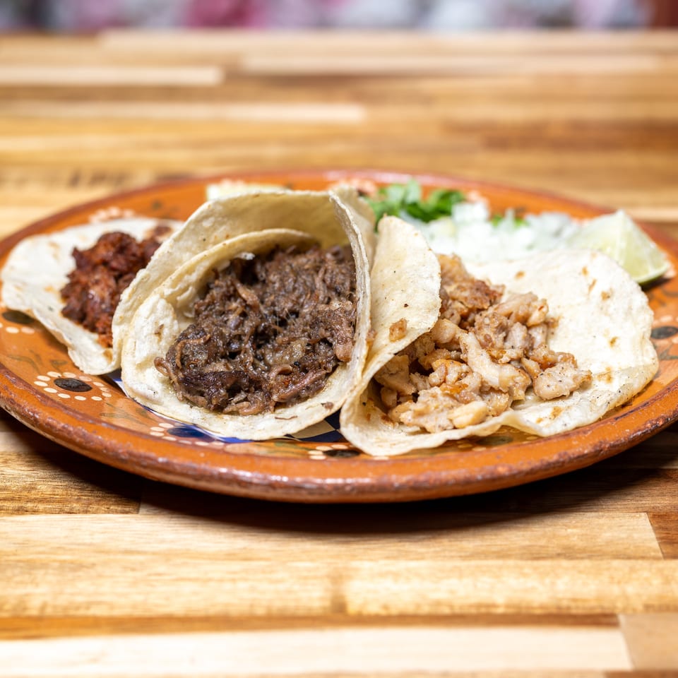 Three tacos with different meat fillings, cilantro, onions, and lime on a decorative plate.