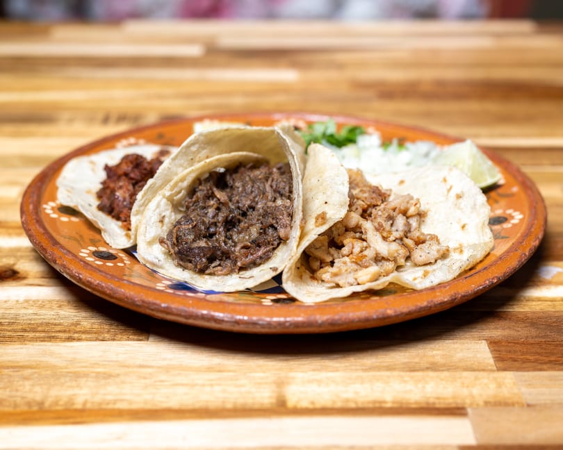 Three tacos with different meat fillings, cilantro, onions, and lime on a decorative plate.
