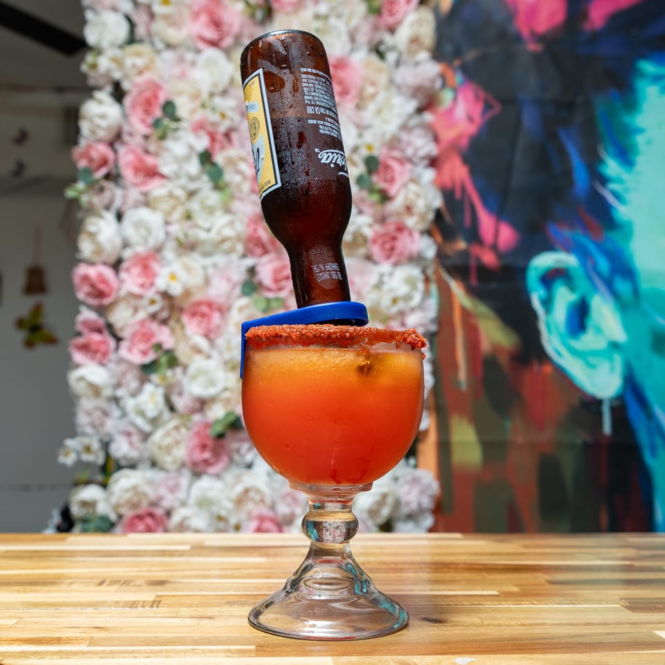 A michelada with a beer bottle inverted into it, on a wooden table with a floral wall.