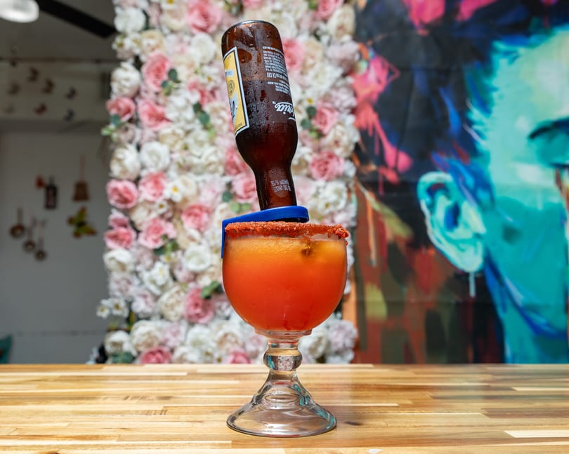 A michelada with a beer bottle inverted into it, on a wooden table with a floral wall.