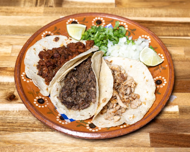 Three street tacos with different meat fillings, cilantro, onions, and lime wedges on a decorative plate.
