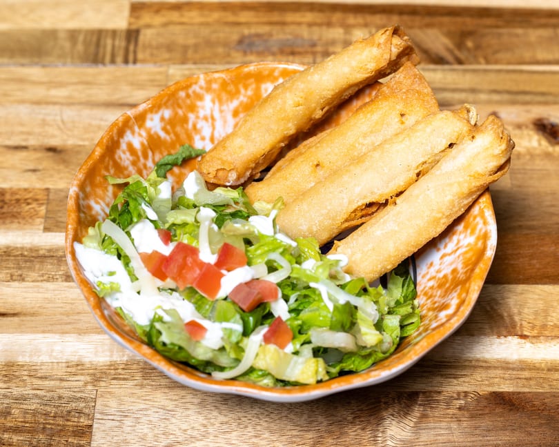 A bowl of golden taquitos with shredded lettuce, diced tomatoes, and sour cream on a wooden table.
