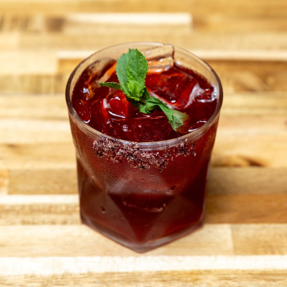 A vibrant red cocktail with ice, garnished with mint and a sugar-rimmed glass on a wooden table.