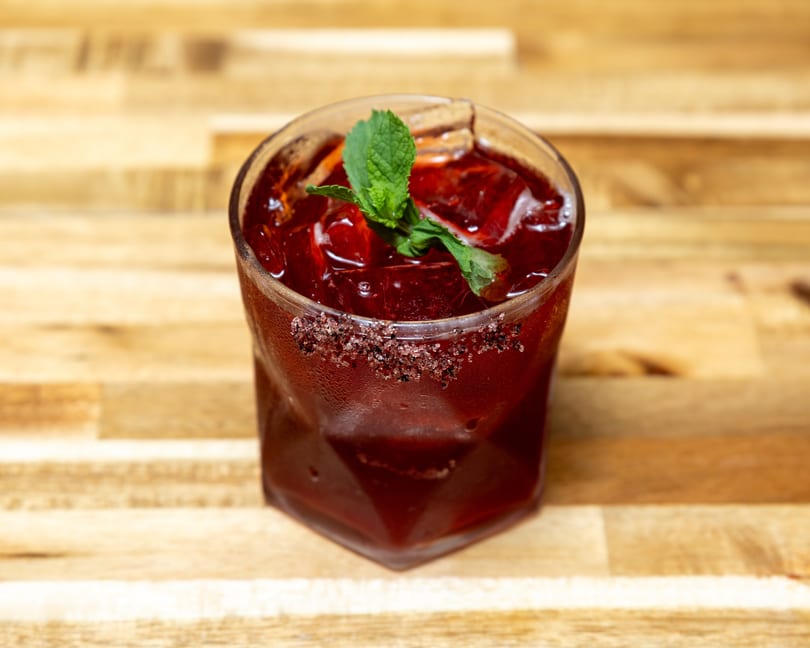 A vibrant red cocktail with ice, garnished with mint and a sugar-rimmed glass on a wooden table.