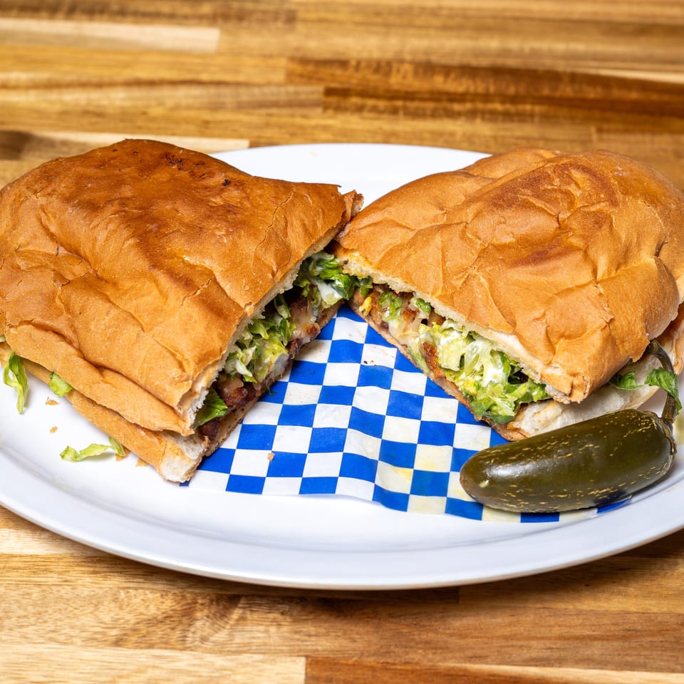 A large sandwich, cut in half, filled with meat and lettuce, served with a pickled jalapeño.