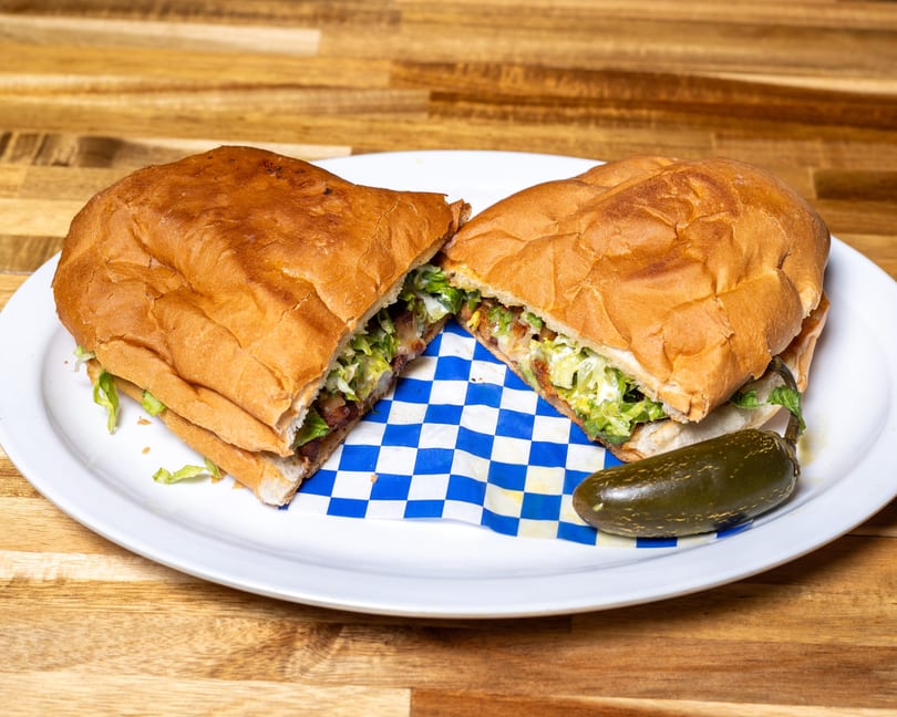 A large sandwich, cut in half, filled with meat and lettuce, served with a pickled jalapeño.