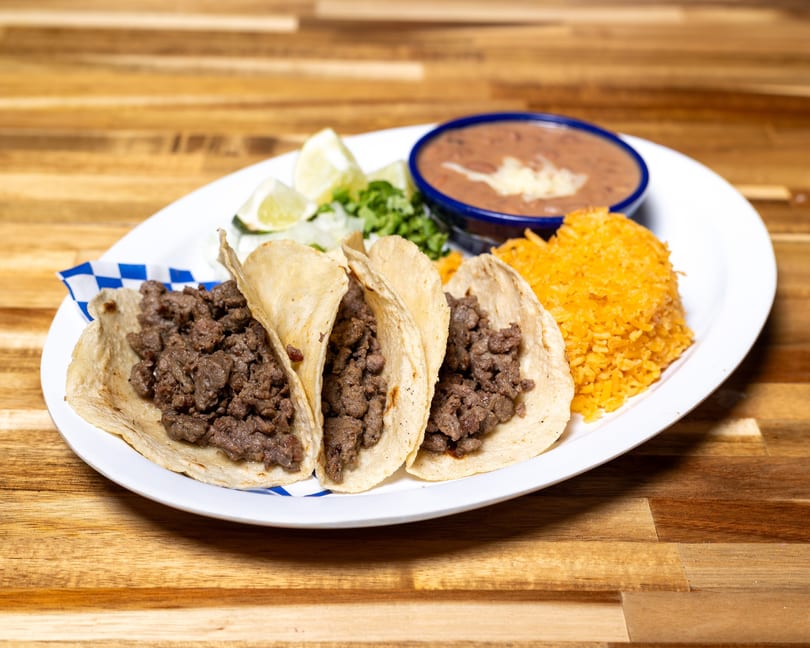 Three beef tacos with lime, cilantro, refried beans, and orange rice on a white plate.