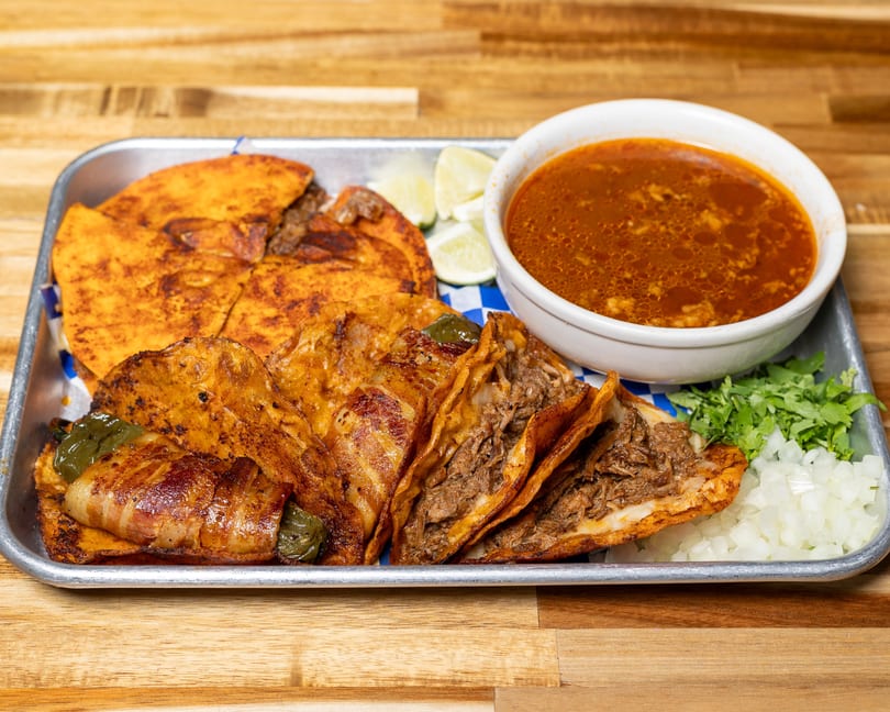 A metal tray with birria tacos, a bacon-wrapped jalapeño, lime wedges, and a bowl of consommé.