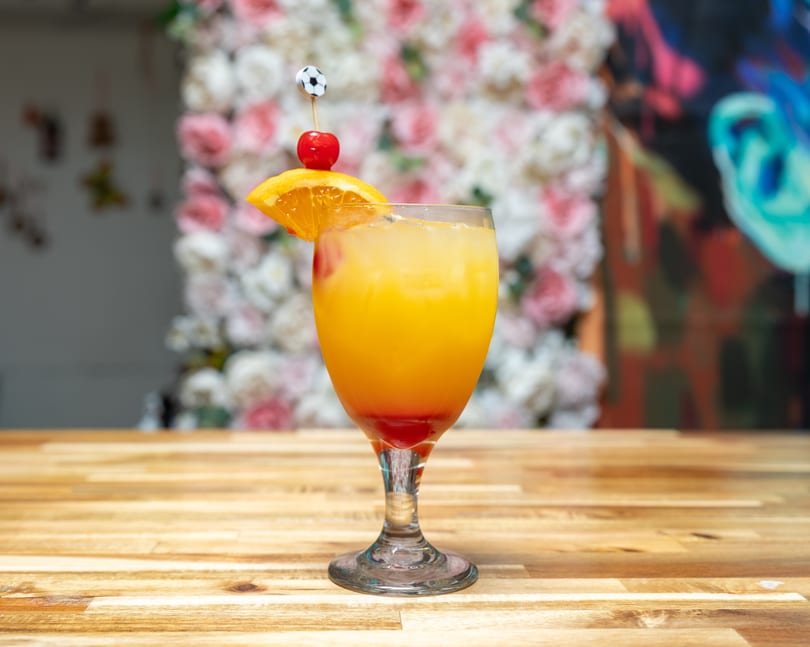 Tropical cocktail with orange, cherry, and soccer ball pick on a wooden table with floral background.