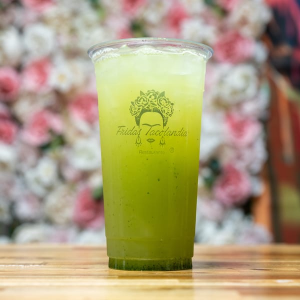 Green drink in a clear plastic cup with the "Fridas Tacolandia" logo on a wooden table.