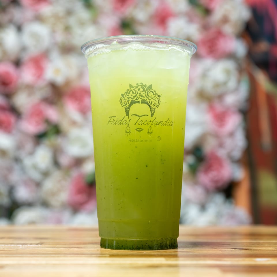 Green drink in a clear plastic cup with the "Fridas Tacolandia" logo on a wooden table.