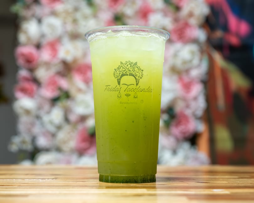 Green drink in a clear plastic cup with the "Fridas Tacolandia" logo on a wooden table.