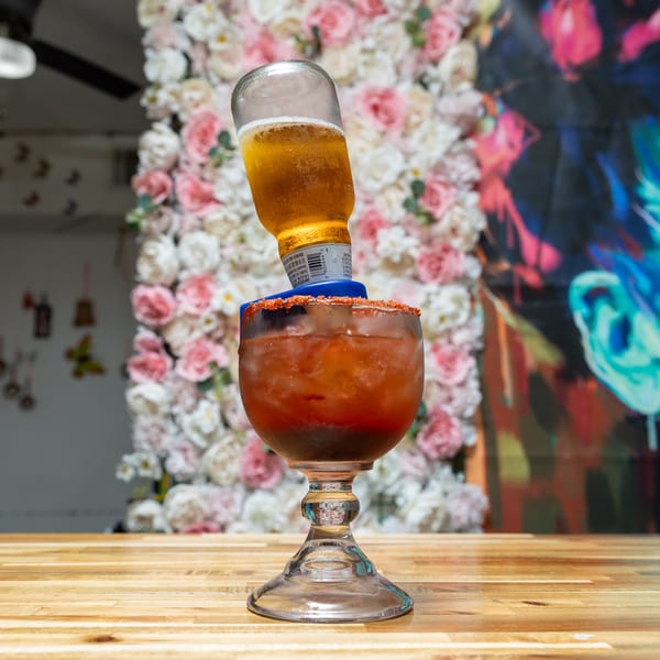 Michelada with an inverted beer bottle, chili-lime rim, on a wooden table with a floral wall.