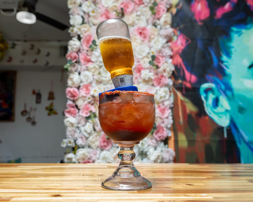 Michelada with an inverted beer bottle, chili-lime rim, on a wooden table with a floral wall.