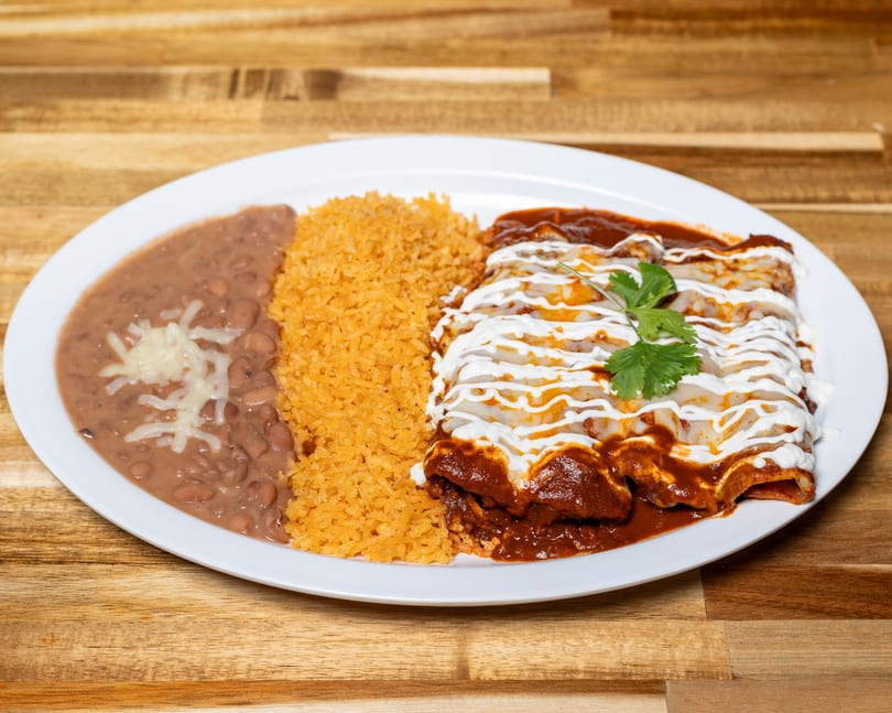 A plate of enchiladas with red sauce, sour cream, rice, and refried beans with cheese.