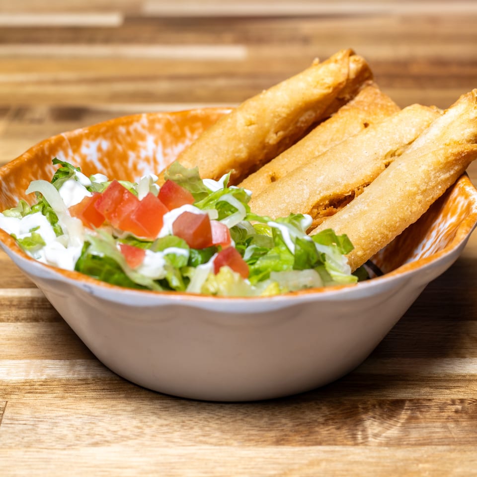 Taquitos with shredded lettuce, diced tomatoes, and sour cream in a white and orange bowl.