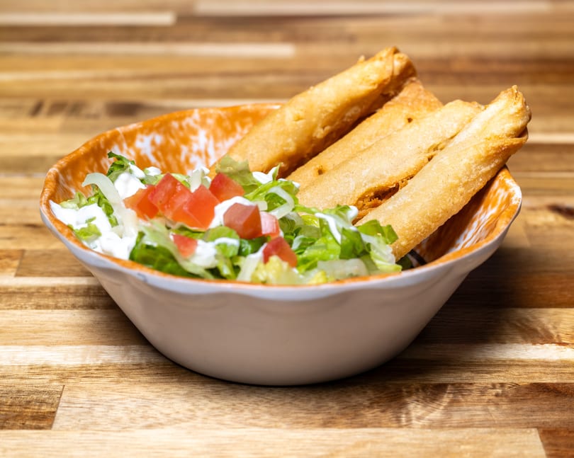 Taquitos with shredded lettuce, diced tomatoes, and sour cream in a white and orange bowl.