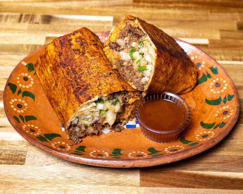 Two halves of a large, grilled burrito filled with meat, rice, and onions, served with dipping sauce.
