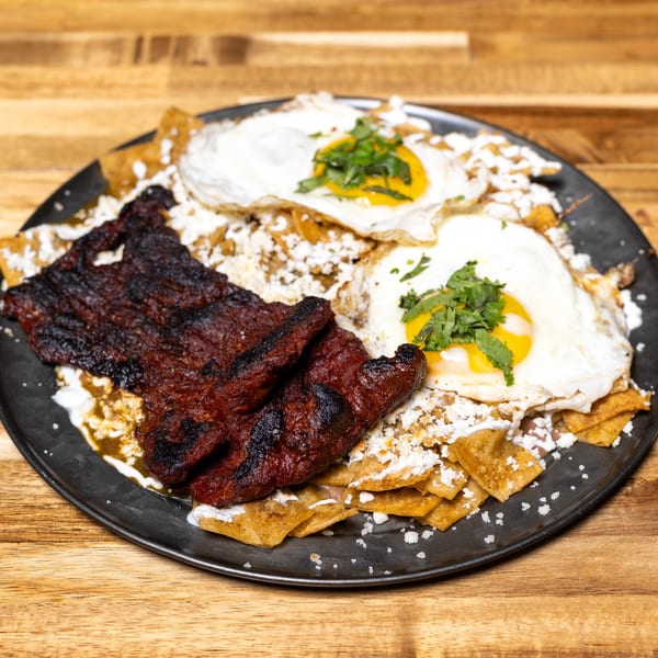 Chilaquiles with grilled steak, two fried eggs, cotija cheese, and cilantro on a dark plate.
