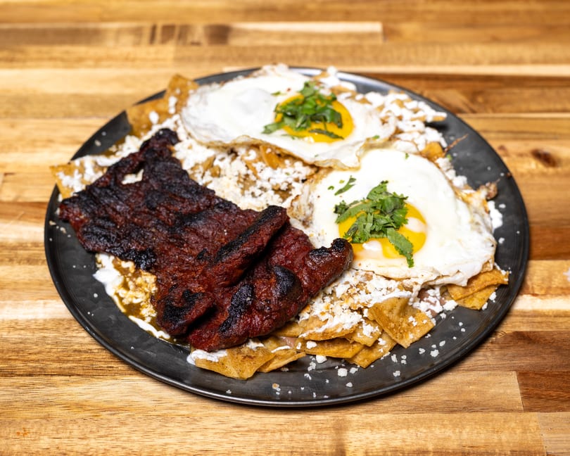 Chilaquiles with grilled steak, two fried eggs, cotija cheese, and cilantro on a dark plate.