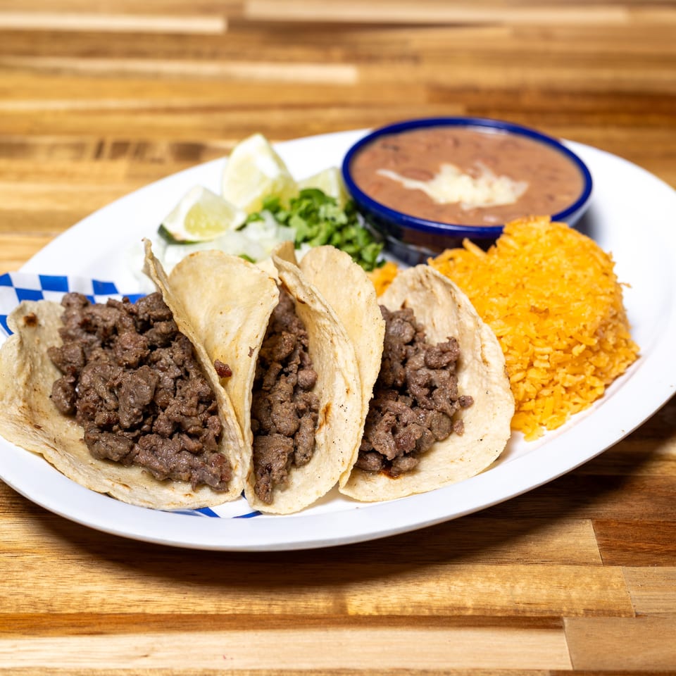Three beef tacos with lime, cilantro, refried beans, and orange rice on a white plate.