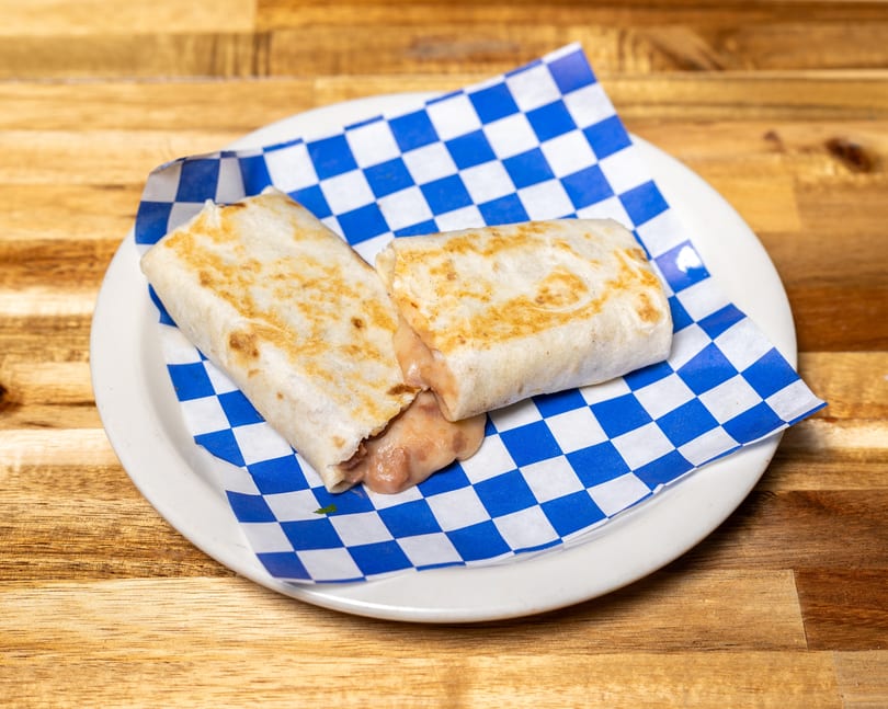 Two golden-brown burritos with melted cheese and beans spilling out, on blue and white checkered paper.