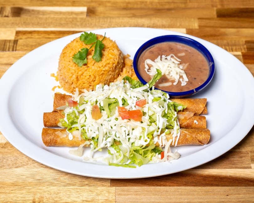 A plate of Mexican food with taquitos, rice, and refried beans on a wooden table.